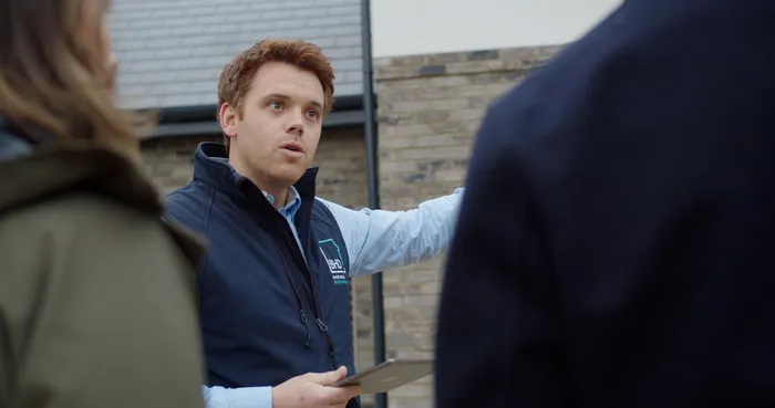 A young man showing two customers the outside of a newly constructed house.