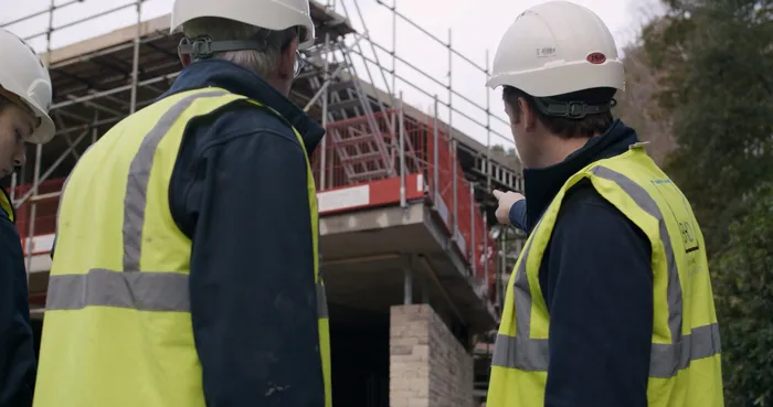 Two builders in high visibility jackets and hard hats look at a house being built.