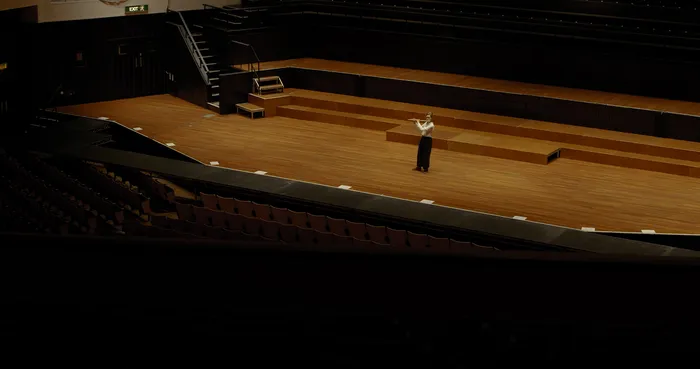 A woman standing alone on a wooden theatre stage.