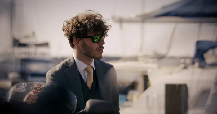 Young man wearing a formal dark grey suit and green polarised sunglasses looking into the distance.