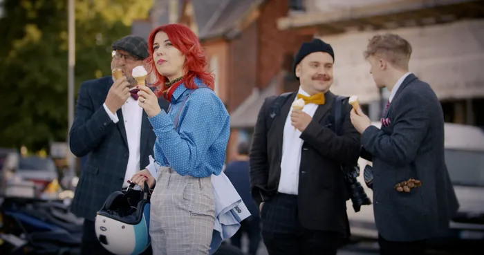 Three young men in suits and a young lady with bright red hair all stand together eating ice cream.